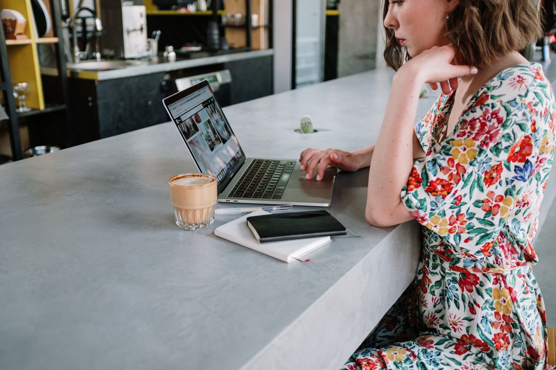 Young woman in a floral print dress working on a laptop in a modern café setting.